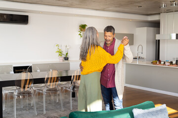A biracial mature couple is dancing together in their living room, at home