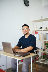 Portrait of smiling Asian man working on laptop at home office. Remote job concept.