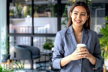 Young happy Asian businesswoman in a suit holding a cup of delivery coffee, standing confident in...