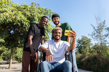 Young black man in a wheelchair using phone taking selfie together with male friends outdoors.