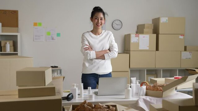 Confident young Asian woman retail seller, entrepreneur, online store dropshipping small business owner looking at camera standing in delivery shipping warehouse with parcel boxes.