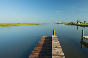 Fototapeta premium A wooden pier extends into a calm body of water under a clear blue sky at the Blue Water RV Beach Resort, Surfside Beach, Texas. The tranquil water seems to merge with the blue summer sky.