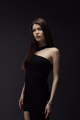 Fototapeta premium A poised young woman in a stylish black dress stands against a dark backdrop, exuding elegance and confidence with a hint of mystery. Perfect for fashion and lifestyle themes.