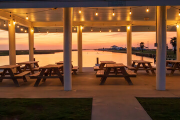 Picnic tables are neatly arranged under a covered pavilion adorned with hanging lights, providing a prime spot to enjoy the serene sunset over the calm waters in Freeport Texas.