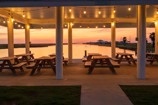 Picnic tables are neatly arranged under a covered pavilion adorned with hanging lights, providing a prime spot to enjoy the serene sunset over the calm waters in Freeport Texas.
