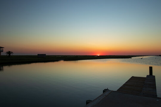 A serene sunset casts a warm glow over a calm body of water at Blue Water RV Resort in Freeport, Texas. A wooden pier extends into the water on the right, while the sun dips just below the horizon.