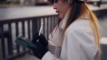 Asian woman listening to the music on earphones and working on digital tablet by the river in Tokyo city, Japan. Attractive girl freelance work anywhere with using gadget device and online network. - Powered by Adobe