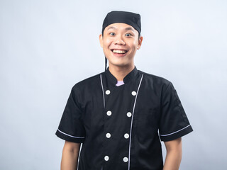 Smiling young man in a black chef uniform and chef hat, standing against a plain light gray background.