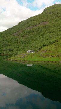 A serene video showing vehicles parked by a mountain lake, with a white van, camper, and RV. The stunning scenery includes a mountain reflected in the calm water, Norweigan Fjords Stavbergvatnet Urke
