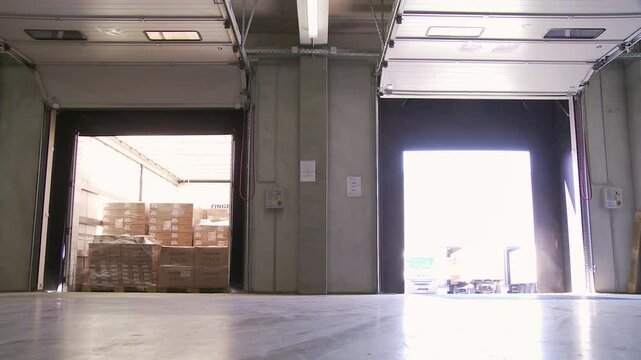 Inside a brightly lit warehouse, sunlight streams through open dock doors revealing stacked cargo boxes