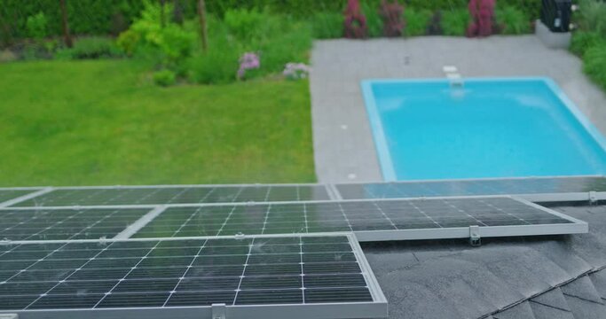 Rain pouring on solar panels installed on a roof with a swimming pool below.