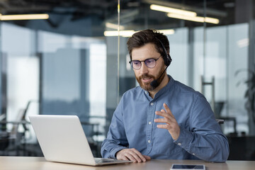 A serious and focused young man businessman conducts a business meeting via video call, sits in the office in a headset in front of a laptop and explains, gesturing with his hands