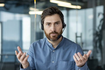 Close-up photo of a young man in a headset and a blue shirt sitting at a desk in an office in front...
