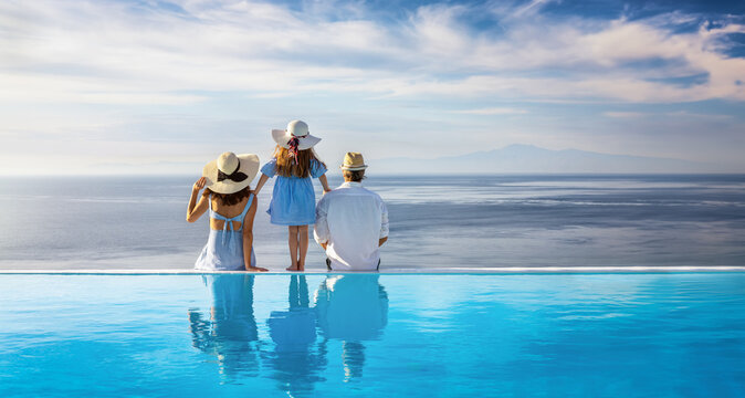 A happy family on summer holidays sits at the edge of a swimming pool and enjoys the beautiful view over the sea