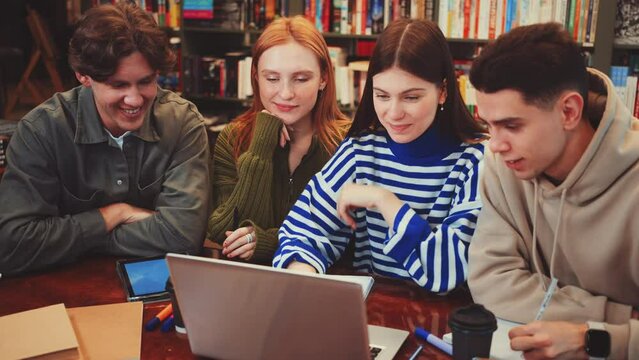 Group of students collaborating in library, gathered around laptop at table. They focus on screen, discussing project. Bookshelves in background. Concept of teamwork and academic collaboration