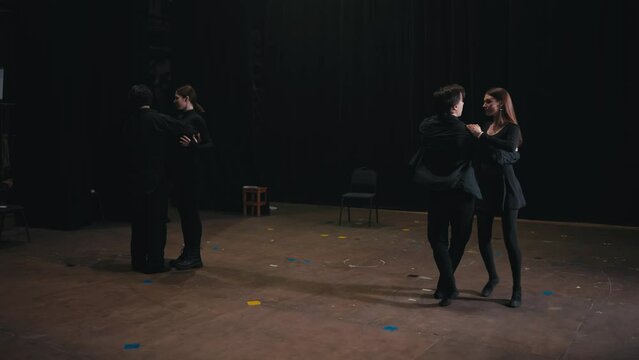 Two pairs of confident actors in black suits dance on stage during their rehearsal for a play in the theater. Careful preparation of actors in black suits before performing and dancing in the theater