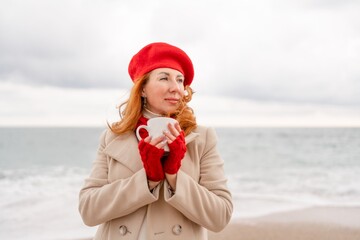 Obraz premium Woman beach in red beret scarf and mitts holding a cup of tea in his hands. Depicting beach relaxation and cozy attire. Walks by the sea
