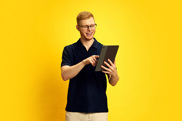 Smiling young redhead man in glasses looking at tablet and pointing with finger, using online services against bright yellow background. Concept of human emotions, facial expression, lifestyle