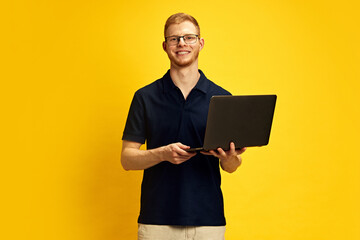 Confident smiling young man in glasses holding laptop against bright yellow background. Business,...