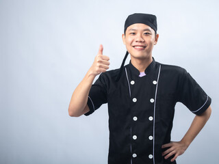 Smiling young man in a black chef uniform, giving a thumbs-up gesture against a plain light gray background. The man exudes confidence and positivity,