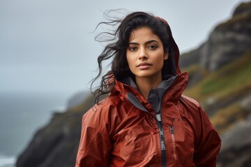 Portrait of a satisfied indian woman in her 20s wearing a functional windbreaker over dramatic coastal cliff background