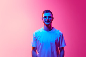 Portrait of young man in casual white t-shirt and glasses posing, looking at camera against pink background in neon light. Concept of human emotions, facial expression, lifestyle
