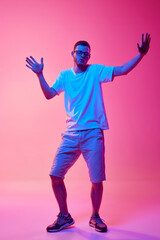 Full-length image of young man in glasses and casual clothes cheerfully dancing and having fun against pink background in neon light. Concept of human emotions, facial expression, lifestyle