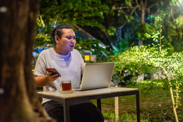 Young Asian man, digital nomad working remotely from cafe, using laptop and smartphone. young Asian men are hanging out in a cafe with a nature theme at night