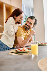 A bearded man and a redhead woman joyfully eating breakfast together in their modern kitchen.