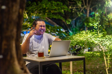 Young Asian man, digital nomad working remotely from cafe, using laptop and smartphone. young Asian men are hanging out in a cafe with a nature theme at night