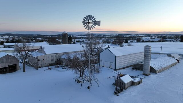 Dawn lights up a snow-covered farm with barns, a silo, and a classic windmill, embodying a serene rural winter scene. Aerial orbit during sunrise.