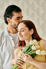 Obraz premium A redhead woman and bearded man embrace and kiss while holding a bouquet of tulips in a modern apartment.