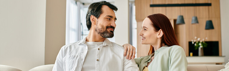 A beautiful adult couple, a redhead woman and a bearded man, sitting on a couch, gazing lovingly at each other in a modern apartment.