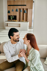 A beautiful adult couple, a redhead woman and bearded man, are sitting together on a cozy couch in a modern apartment.