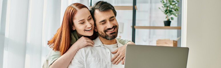 A redheaded woman and a bearded man hugging while enjoying a moment together by looking at a laptop in a modern apartment.