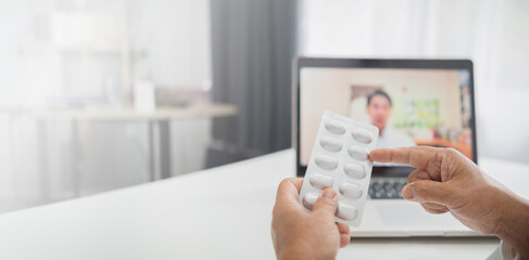 A patient holds a blister pack of pills while consulting a doctor via a video call on a laptop,...