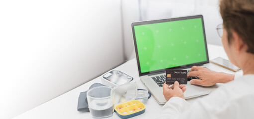 An elderly person uses a credit card for an online transaction on a laptop with medical equipment...
