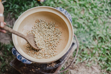 Roasting coffee beans in a ceramic bowl using a wooden spoon outdoors, surrounded by greenery. Ideal for harvest season designs or culinary and cultural events.