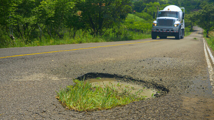 CLOSE UP: Big hole in the asphalt road with approaching lorry in the background