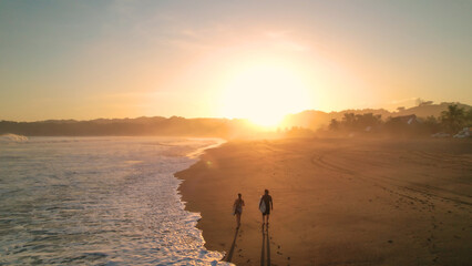 AERIAL Sport travellers walking on beach with surfboards in golden morning light