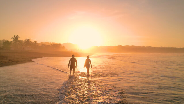 AERIAL: Tidal wave washes on sandy beach and crosses path of two walking surfers