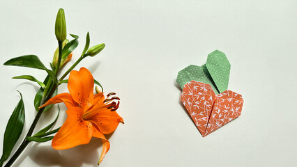 A striking orange lily next to two green and red origami hearts on a white background, perfect for...