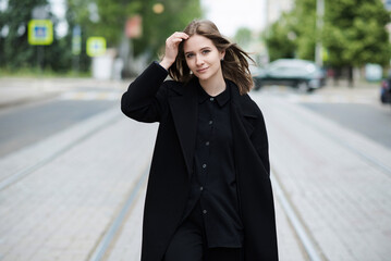 Fototapeta premium Attractive caucasian young woman in black coat walking down city street on warm autumn day. Outdoors portrait. Model in stylish clothes. Lifestyle, fashion concept.