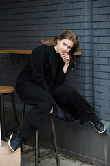 Attractive Caucasian young woman in black coat sitting near cafe while walking along city street on autumn day. Outdoors portrait. Lifestyle, fashion concept. Stylish clothes.