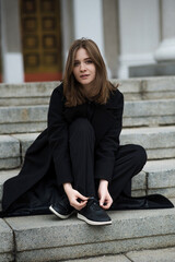 Attractive Caucasian young woman in a black coat sitting on the stair while walking along the city street on autumn day. Outdoors portrait. Travel, rest, fashion, style concept.