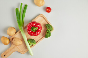 Top view advertising photo, the wooden tray contains a red bell pepper, some pieces of green broccoli, and a leek, featured on white countertop. Beside decorated with some fresh potatoes, copy space