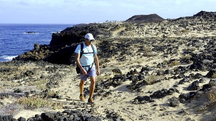 Hiker walks on sandy path between lava stones on the volcanic coast of Lanzarote in the Canary Islands. Healthy summer vacation concept