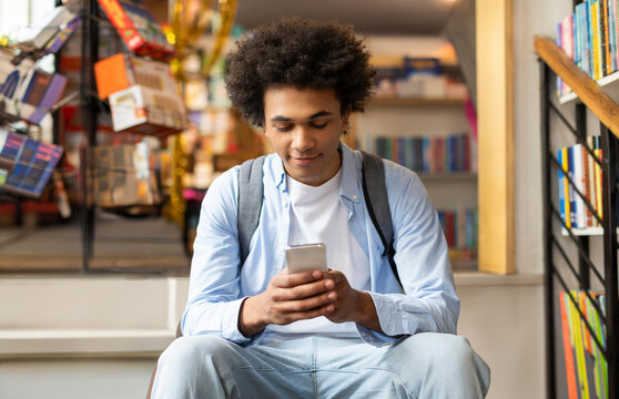 Black male student chatting social media on smartphone, sitting in library after passing the exam in university