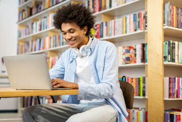 Satisfied Black male student with curly hair, sitting in college library, using laptop and...