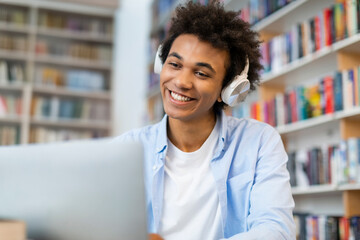 Cheerful Black student man in wireless headphones looking at laptop and smiling, talking on video...